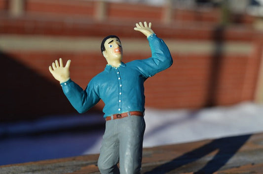 A frightened looking male figurine with his hands up in the air as if he is surprised. He wears a blue shirt and gray pants and there is a red brick wall behind him